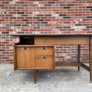 May include: A mid-century modern desk with a wood top and a cabinet with drawers. The desk has a simple design with tapered legs and a small shelf above the cabinet.