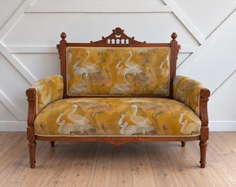 May include: A wooden loveseat featuring a rich brown frame with carved detailing and turned legs. The upholstery is a vibrant mustard yellow fabric decorated with a repeating pattern of white and light brown cranes in various poses.