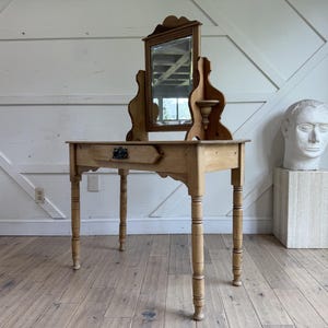 May include: A light brown wooden vanity table featuring a rectangular mirror attached to the back, a single drawer with a dark metal pull, and four turned legs. The mirror is flanked by two small decorative shelves on either side of the frame.