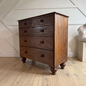 May include: A wooden chest of drawers with five drawers and round knobs. The chest is made of dark wood and has a warm, natural finish. It stands on four rounded feet. A white sculpture sits on a wooden block to the right.