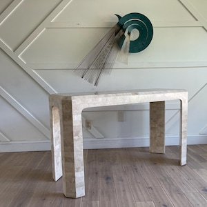 May include: A white, rectangular console table with a unique, geometric design. The table has a smooth, polished surface and is made of a material that resembles marble. The table is standing on a wooden floor.
