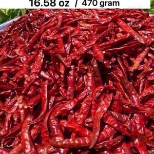 May include: A large pile of stemless dried red Jinda dang chilies. The text at the top reads: พริกจินดาแดง แห้ง เด็ดก้าน Dried jinda dang chilies | 16.58 oz / 470 gram. The chilies are deep red, elongated, and have a wrinkled, dried texture.