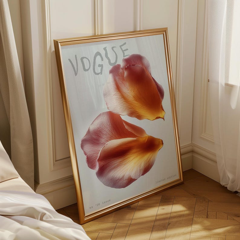May include: A framed print featuring the word "VOGUE" in light gray, with two large, overlapping flower petals in shades of orange and red. The print is in a gold frame and leans against a white wall.