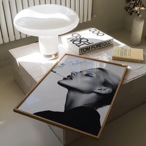 May include: A white mushroom-shaped table lamp with a gold base sits on a white tiled surface. A framed black and white portrait of a woman smoking a cigarette is displayed nearby, along with two Tom Ford books and a book titled "Gentle Living".