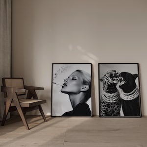 May include: Two framed black and white art prints. One features a woman smoking, the other shows two big cats wearing pearl necklaces. The prints are displayed in a room with a wooden chair.