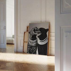 May include: A framed black and white print featuring two big cats, a leopard and a panther, adorned with pearl necklaces. The leopard has a spotted coat, while the panther is solid black. The artwork is leaning against a wall.