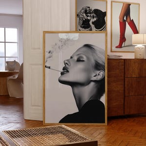 May include: Black and white framed print of a woman exhaling cigarette smoke. The image is in a wooden frame and is leaning against a wall. Other framed art pieces are visible in the background.
