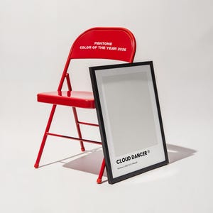 May include: A red metal folding chair with the text "PANTONE COLOR OF THE YEAR 2026" on the backrest. A black framed print with the text "CLOUD DANCER" is leaning against the chair.