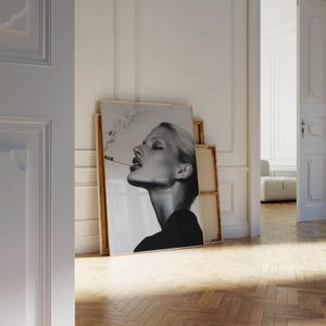 May include: Black and white framed print of a woman smoking a cigarette. The woman has blonde hair and is wearing a black top. The print is leaning against a wall in a room with a wooden floor.