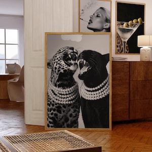 May include: A framed black and white print featuring a leopard and a panther, both wearing pearl necklaces and roaring. Additional framed prints include a woman smoking and a martini glass with olives. The artwork is displayed in a room with wooden furniture.