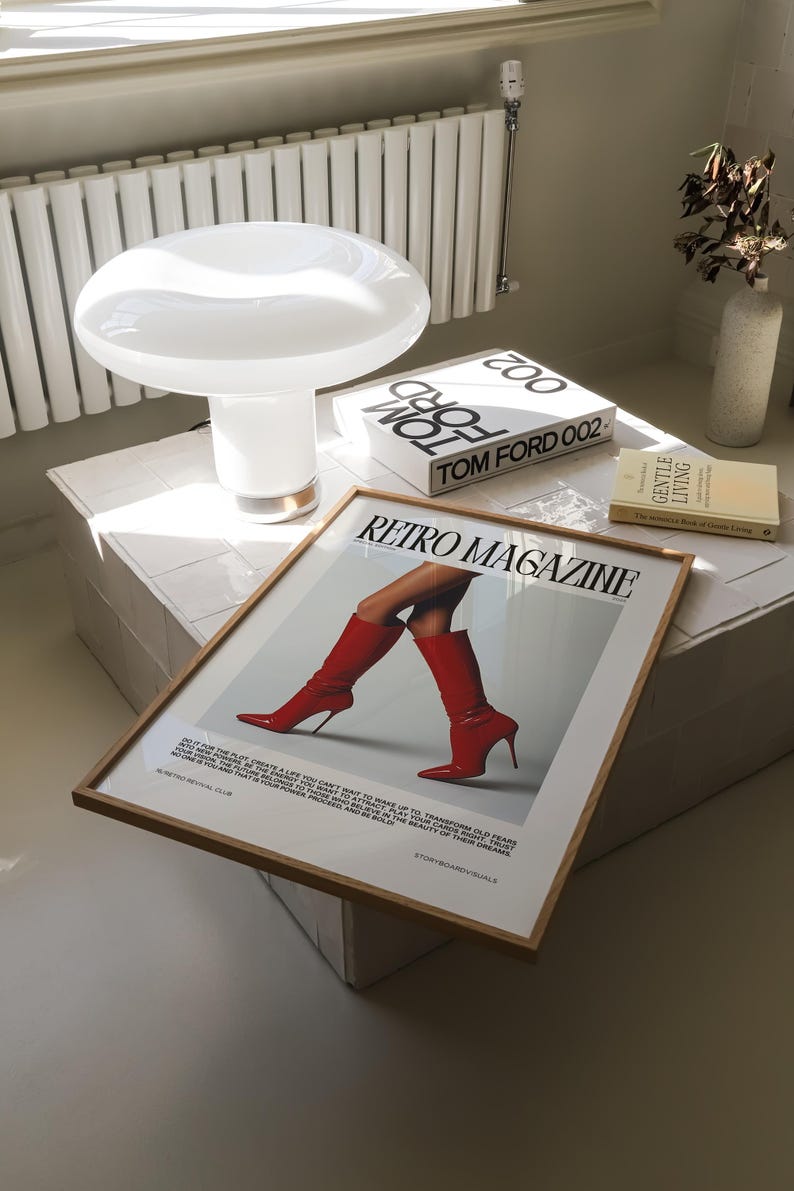 May include: A white mushroom-shaped lamp with a gold base sits on a white tiled surface, next to a framed "Retro Magazine" print featuring red boots. Also present are two Tom Ford books and a vase with dried flowers.