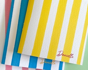 May include: Three notebooks with striped covers in pink, blue, and yellow with white stripes. Each notebook has the name "Danielle" written in a script font. The notebooks are stacked on a light green surface.