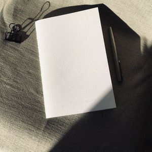 May include: A white rectangular notebook with a textured cover featuring the embossed text wildcardinland studio in the bottom right corner, accompanied by a black metal binder clip and a slim black pen positioned on a neutral fabric surface.