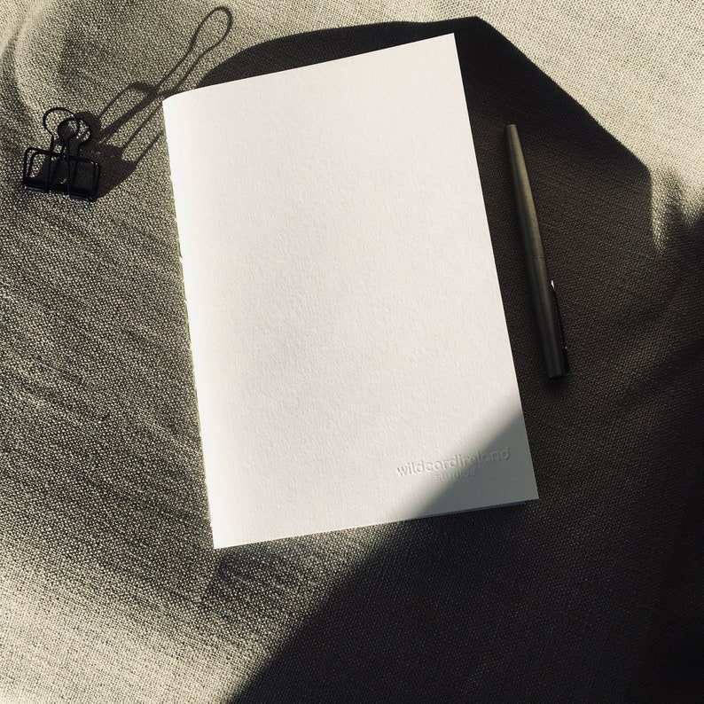 May include: A white rectangular notebook with a textured cover featuring the embossed text wildcardinland studio in the bottom right corner, accompanied by a black metal binder clip and a slim black pen positioned on a neutral fabric surface.