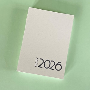 Può includere: Un'agenda di colore chiaro per l'anno 2026, con la parola "DIARY" stampata verticalmente e il numero "2026" in grassetto. L'agenda è posta su uno sfondo verde chiaro. Un quaderno per pianificare.
