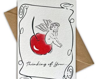 May include: A white greeting card featuring a black ink illustration of a cherub sitting on a bright red cherry. The card has a decorative black border and the text Thinking of You... written in black script at the bottom, paired with a brown envelope.