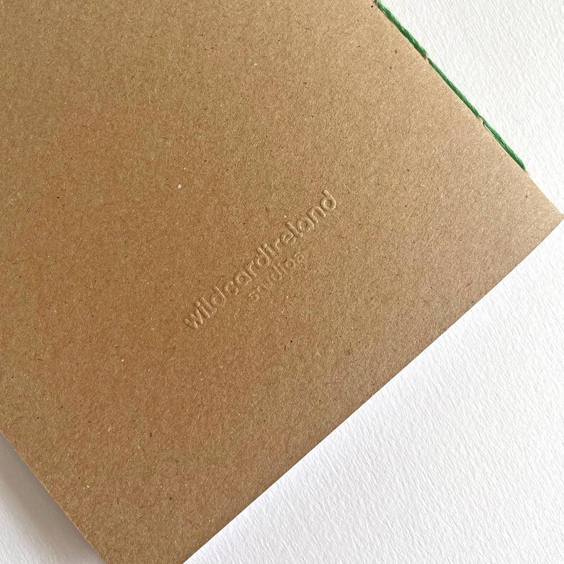 May include: A rectangular brown kraft paper card with a debossed logo that reads wildcardireland studios. A thin green thread is visible along the top right edge of the card, which features a smooth, matte texture and a clean, minimalist design.