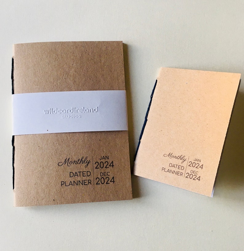 May include: Two tan paper planners with black stitched spines. The larger planner features a white paper belly band embossed with the text wildcardireland studios. Both planners are printed with the text Monthly DATED PLANNER JAN 2024 DEC 2024.