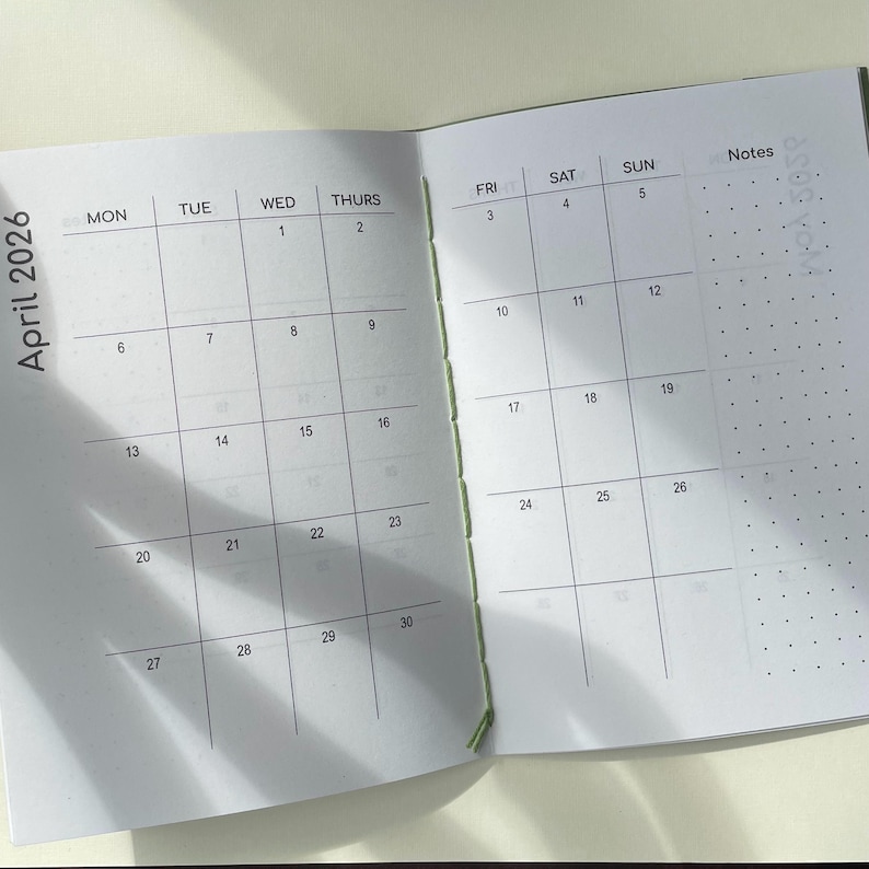 May include: An open planner showing the month of April 2026 with a grid layout for days Monday through Sunday. The pages feature black printed text, a dotted notes section on the right, and a light green thread binding visible along the center spine.