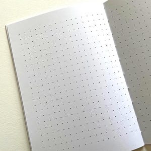 May include: An open notebook with white pages featuring a grid of small, evenly spaced black dots. The paper is bound along the right side with visible stitching, creating a clean layout for writing, sketching, or organizing notes on the page.