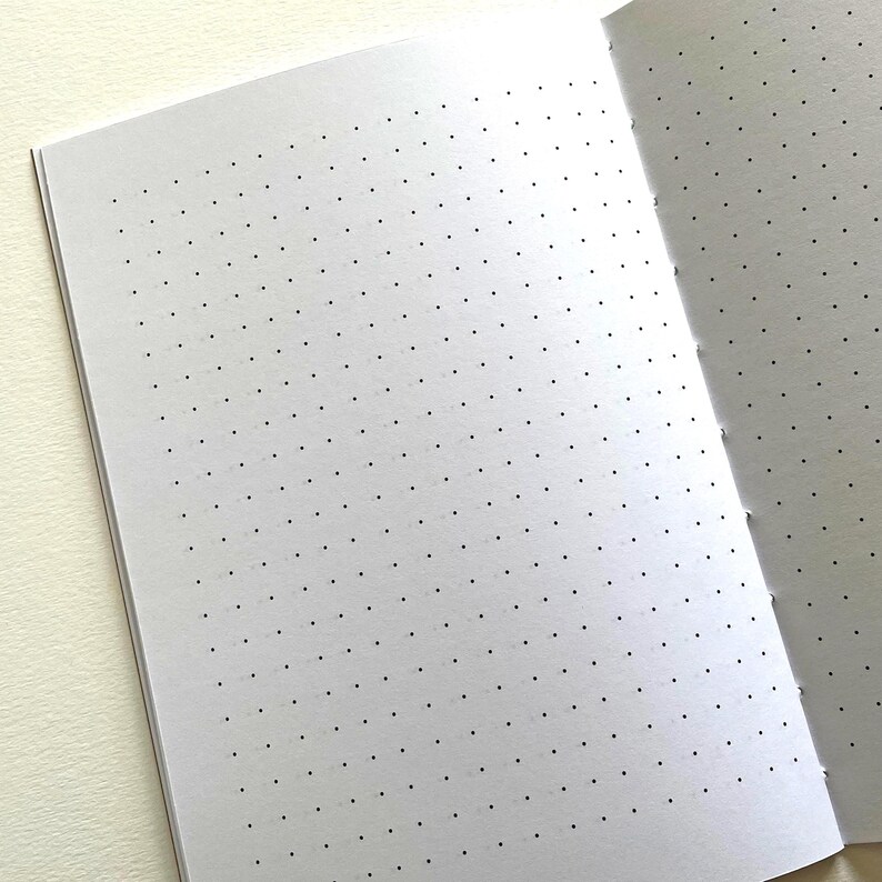 May include: An open notebook with white pages featuring a grid of small, evenly spaced black dots. The paper is bound along the right side with visible stitching, creating a clean layout for writing, sketching, or organizing notes on the page.