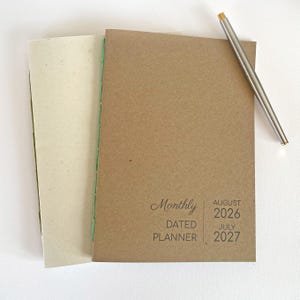 May include: A brown paper-covered monthly dated planner with green thread binding, featuring the text Monthly DATED PLANNER AUGUST 2026 JULY 2027 on the front cover, accompanied by a slim silver metal pen with a gold-colored tip.