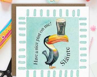 May include: A square greeting card featuring a watercolour illustration of a toucan balancing a pint of Guinness on its beak. The text reads Have a nice pint on me! and Sláinte. The card has a light blue background with a dashed border pattern.