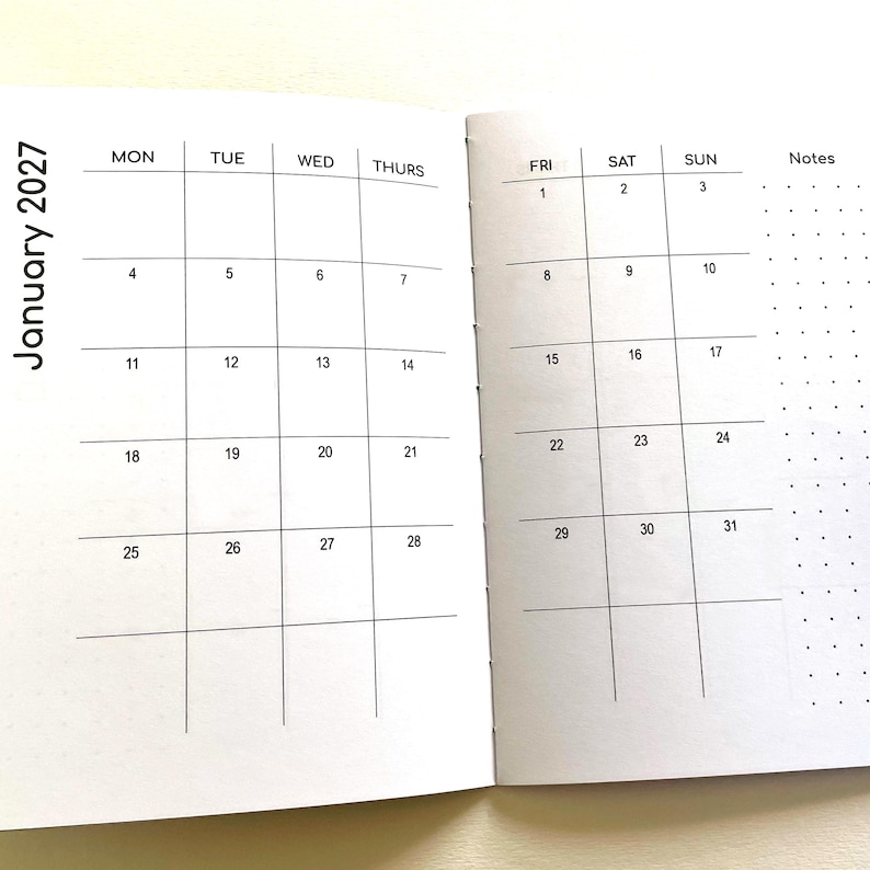 May include: An open planner showing a monthly calendar for January 2027. The left page displays days Monday through Thursday, while the right page shows Friday through Sunday with a dotted notes column. The layout features black grid lines.