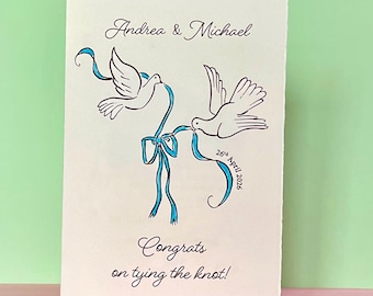 May include: A cream wedding card featuring two doves with a blue ribbon design. The card is inscribed with "Andrea & Michael" and "Congrats on tying the knot!". The date "26th April 2026" is also included in the design.