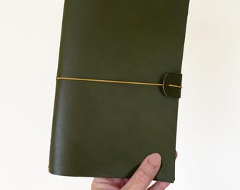 May include: An olive green leather journal with a matching closure tab and a tan elastic band. The journal is held in a hand against a white background. The cover is smooth and the edges are clean.
