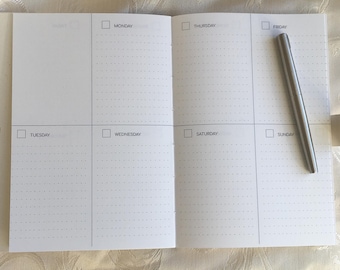 May include: An open weekly planner with white pages featuring a grid of six daily sections labelled Monday through Sunday, each with a checkbox and dot grid paper. A sleek silver metal pen rests diagonally across the Sunday section of the planner.