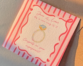 May include: A square engagement card with a watercolour illustration of a ring. The card has a pink and white striped border and the text "Jane & Liam has a nice ring to it. Congrats on your engagement!"