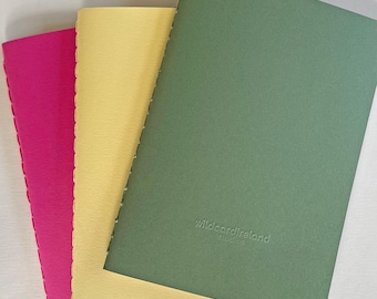 May include: Three notebooks in pink, yellow, and green are stacked. The notebooks have a stitched binding and a textured cover. The green notebook has the text "wildcardireland studios" embossed on the cover.