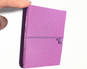 May include: A small, rectangular notebook with a solid purple fabric cover. The notebook is held closed by a thin, matching purple elastic cord that wraps horizontally around the centre and is secured with a small knot on the front cover.