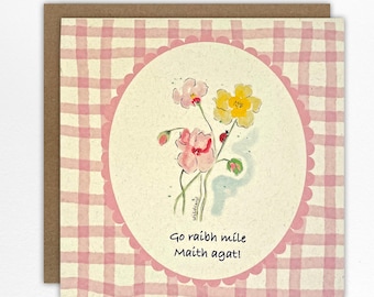 May include: A greeting card with a watercolor floral design. The card features pink and yellow flowers with green leaves, set against a cream-coloured oval. The background has a pink and white gingham pattern, with the text "Go raibh míle Maith agat!"