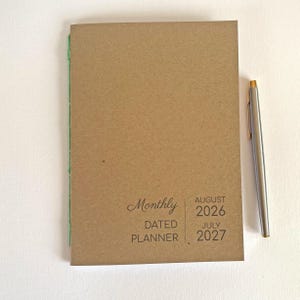 May include: A tan monthly dated planner with green spine stitching and a silver pen. The cover features the text Monthly DATED PLANNER AUGUST 2026 JULY 2027. The planner has a simple, rectangular design with a clean, neutral-toned paper cover.