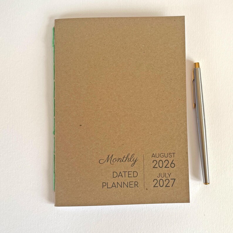 May include: A tan monthly dated planner with green spine stitching and a silver pen. The cover features the text Monthly DATED PLANNER AUGUST 2026 JULY 2027. The planner has a simple, rectangular design with a clean, neutral-toned paper cover.