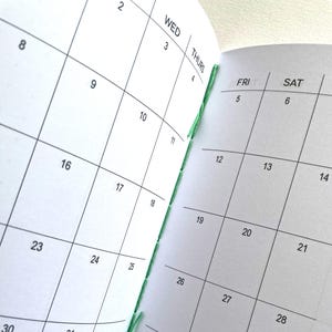 May include: An open planner with white pages and black grid lines showing a monthly calendar layout. The days of the week WED, THURS, FRI, and SAT are printed at the top of the columns. The binding features visible green thread stitching.