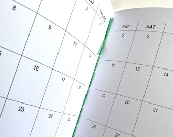 May include: An open planner with white pages and black grid lines showing a monthly calendar layout. The days of the week WED, THURS, FRI, and SAT are printed at the top of the columns. The binding features visible green thread stitching.