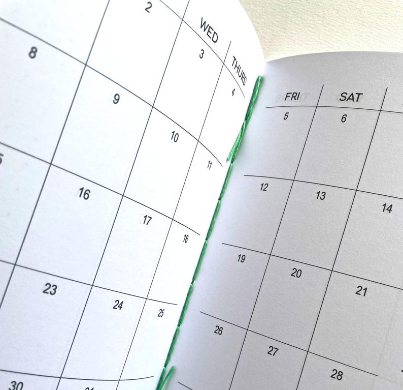 May include: An open planner with white pages and black grid lines showing a monthly calendar layout. The days of the week WED, THURS, FRI, and SAT are printed at the top of the columns. The binding features visible green thread stitching.