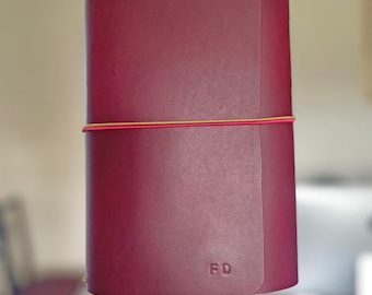 May include: A burgundy leather journal with a wraparound closure. The journal is secured with a pink and gold elastic band. The initials "FD" are embossed on the lower right corner. The journal is held by a person.