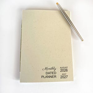 May include: A light beige monthly dated planner with a speckled texture. The cover features black text reading Monthly DATED PLANNER AUGUST 2026 JULY 2027. A slim silver metal pen with a gold-colored tip rests diagonally across the top right corner.