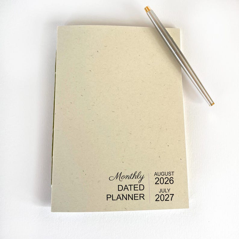 May include: A light beige monthly dated planner with a speckled texture. The cover features black text reading Monthly DATED PLANNER AUGUST 2026 JULY 2027. A slim silver metal pen with a gold-colored tip rests diagonally across the top right corner.