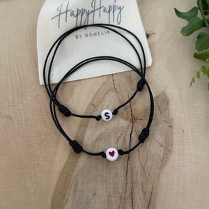 May include: Two black string bracelets with white beads. One bead has the letter "S" and the other has a red heart.
