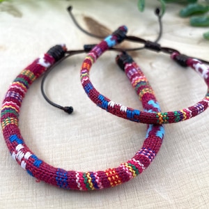 May include: Two colorful woven bracelets with a red, blue, white, yellow, and green pattern. The bracelets are tied with a black cord.