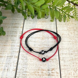 May include: Two adjustable string bracelets. One is red, the other is black. Each bracelet has a black bead with a white letter. The red bracelet has the letter "A", and the black bracelet has the letter "M".