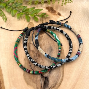 May include: Three colorful woven bracelets with black string ties. The bracelets are made of different patterns and colors, including green, red, black, white, and blue.