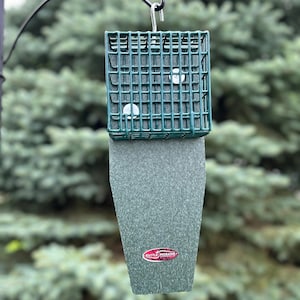 May include: A green bird feeder with a square, grid-like top and a textured, rectangular bottom. The feeder is suspended from a black metal hook against a backdrop of green foliage. The Kettle Moraine logo is visible.