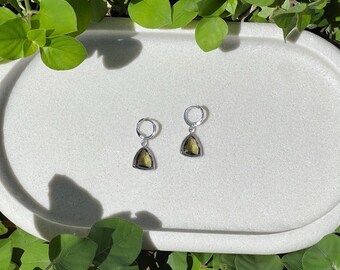 May include: A pair of silver hoop earrings with triangular, olive-green gemstones. The earrings are displayed on a white, oval-shaped tray, surrounded by green foliage. The gemstones have a faceted appearance, catching the light.
