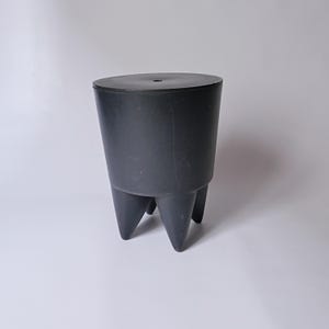 May include: A black, three-legged stool with a round top. The stool has a cylindrical body that tapers slightly towards the bottom. The legs are triangular and extend downwards. The stool is made of a matte material.